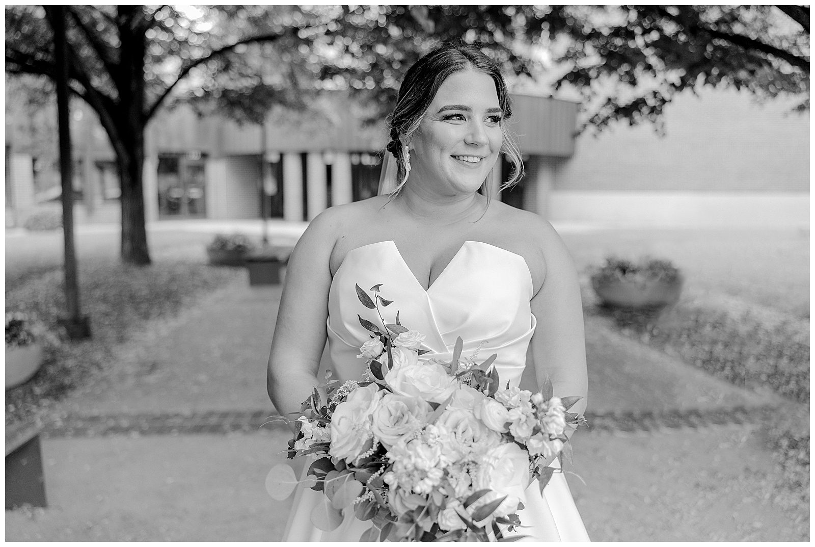 black and white image of bride in strapless stress, smiling off to the right while holding a bouquet of roses and eucalyptus. 