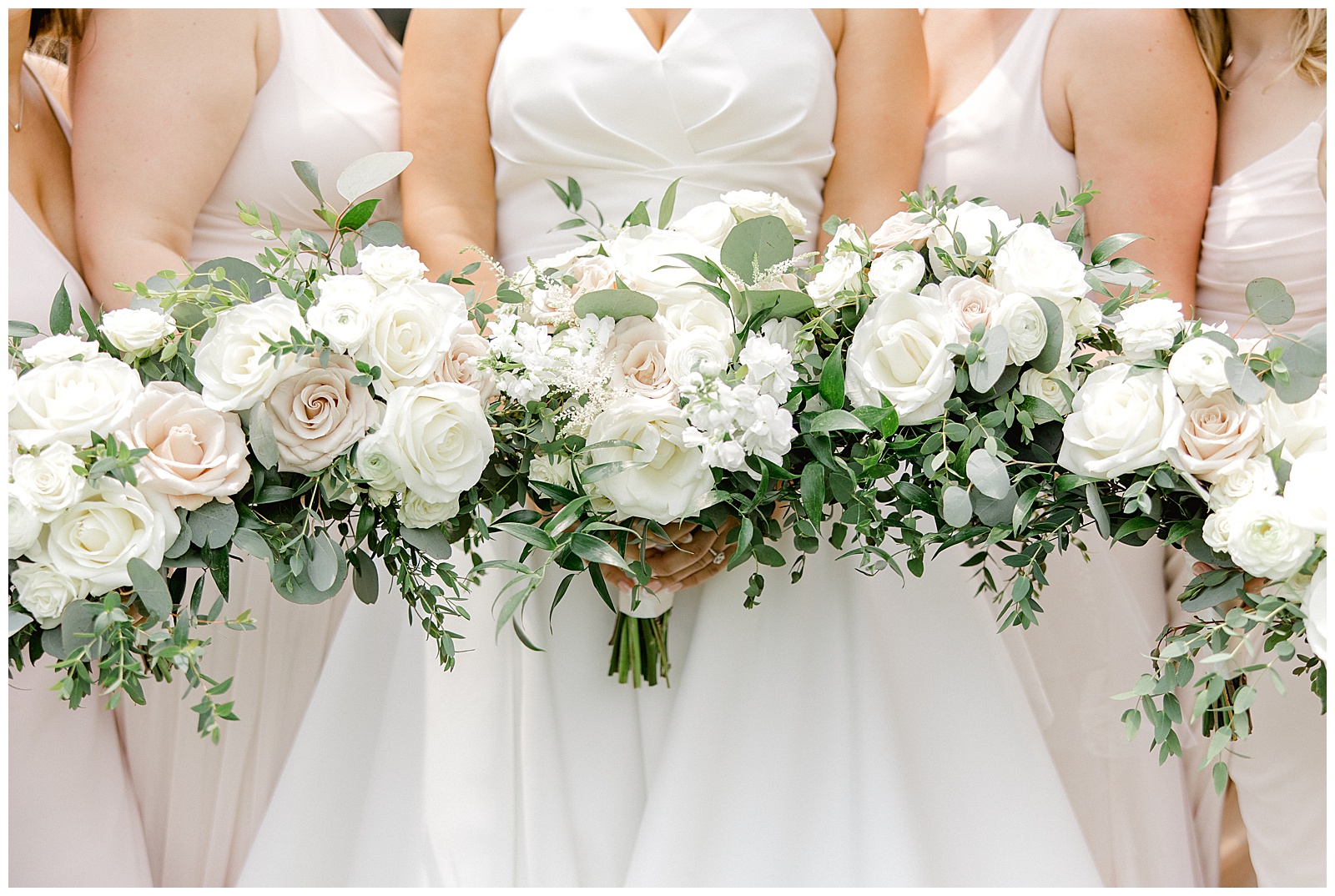 bride in the middle with two ladies on each side of her. faces are not visible. Image is focused on the wedding bouquet consisting of white and blush roses with eucalyptus. 